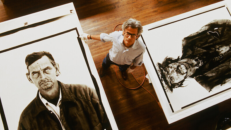 An artist stands between two large black-and-white portraits on tables in a studio with wooden floors, looking up at the camera; the environment is bright and spacious, emphasizing the creative setting and detailed artwork.