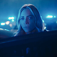 A woman with long blonde hair looks distressed in a nighttime scene with blue emergency lights and blurred police cars in the background, captured through a car window.