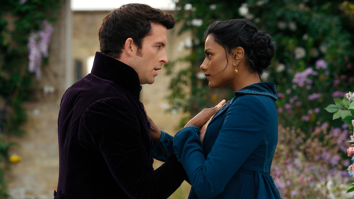 A man and woman in period clothing stand close together, gazing into each other's eyes in an outdoor garden with stone walls and blooming flowers, conveying a dramatic or romantic moment.