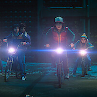 Group of kids riding bicycles at night in an urban or suburban setting, illuminated by their bike headlights, with a mysterious, adventurous atmosphere.