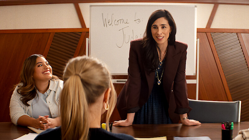 Three women in a meeting room, one standing and smiling while leaning on a table, two seated, with a whiteboard in the background that says "Welcome to". Professional office setting.