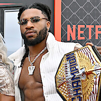 Trick Williams in sunglasses and open white shirt holding WWE United States Championship belt, standing in front of a Netflix display at an event.