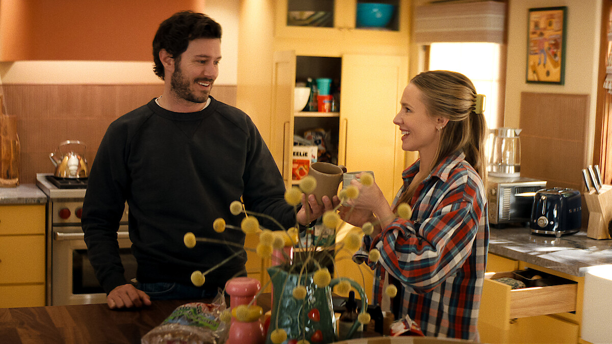 Two people smiling and toasting mugs in a cozy, brightly lit kitchen with yellow cabinets, assorted groceries on the counter, and decorative items in a homey setting.
