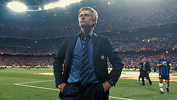 Soccer coach in a suit standing confidently on a stadium field, with players and a large crowd in the background during a nighttime sports event.