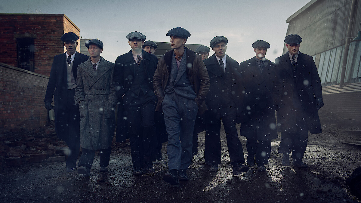 Eight men in vintage suits and flat caps walk together on a wet, gritty street in an industrial urban area, with old brick and metal buildings in the background under a gray, overcast sky.