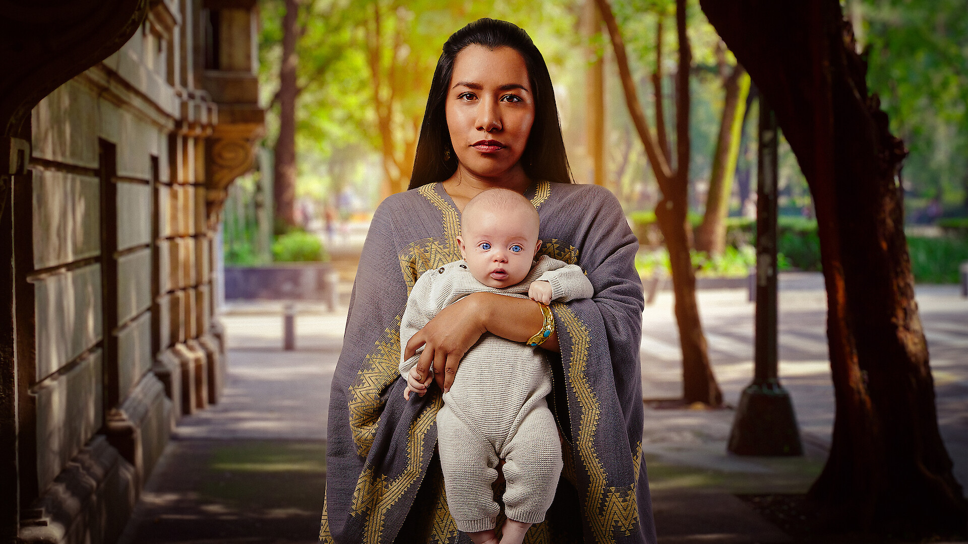 Woman in traditional attire holding a baby, standing on a city street lined with trees and buildings, with soft daylight filtering through the foliage.