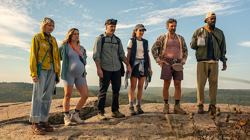 Six hikers stand on a rocky hilltop in casual outdoor gear, overlooking a scenic landscape under a blue sky with light clouds, suggesting a group hiking adventure in nature.