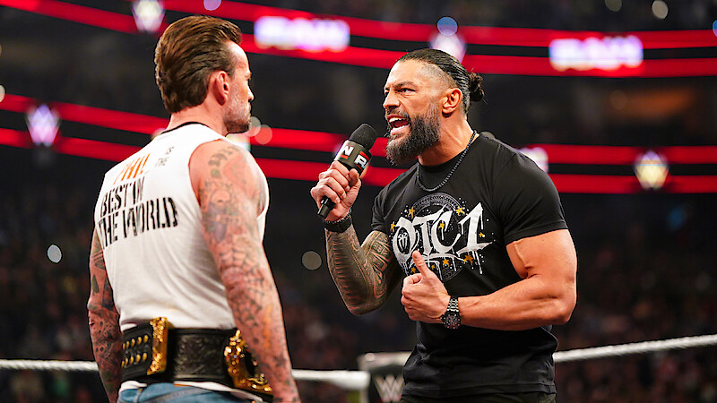 CM Punk and Roman Reigns face off in a crowded arena; one holds a microphone and passionately speaks while the other, with championship belts, listens. Bright lights and audience are visible in the background.