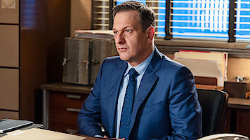 Man in a blue suit sitting at a desk in an office environment, with files, folders, and boxes on wooden furniture, blinds on the window letting in daylight behind him.