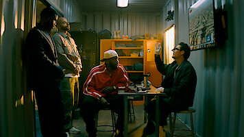 Four men in a dimly lit storage room; three stand while one in glasses gestures, sitting at a table. Shelves, jackets, and a wall map add to the gritty, informal setting.