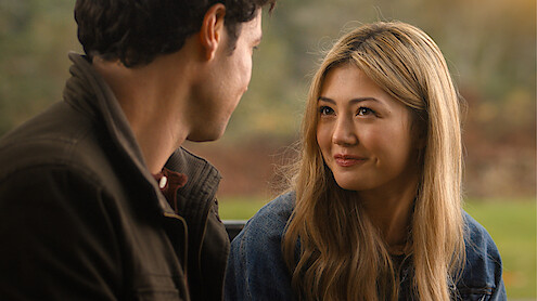 A young woman with long blonde hair smiles warmly at a man while sitting outdoors in a park-like setting with blurred greenery in the background, suggesting a friendly or romantic conversation.