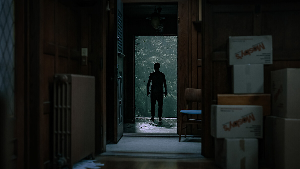 Silhouette of a person standing in a doorway, seen from inside a dimly lit room with moving boxes and wooden paneling, looking out toward a green, misty environment.