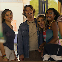 Four young adults smiling and laughing together outdoors in a sunny setting, wearing casual summer clothes, appearing relaxed and happy in a social environment.
