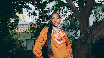 Person standing outdoors in a garden wearing an orange top and patterned scarf, posing confidently with hand on hip, large tree and house with tiled roof in the background, surrounded by greenery.