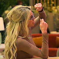 Elissa with long blonde hair and glasses in a beige top gestures with her hands while speaking, sitting indoors on a couch with colorful cushions and greenery in the background.