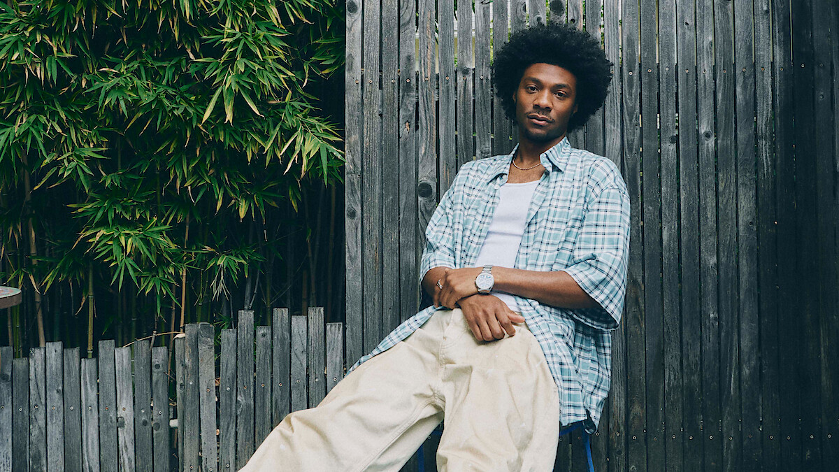 Jonathan Daviss with an afro hairstyle sits casually against a wooden fence outdoors, wearing a plaid shirt over a white t-shirt and cream pants, with green foliage in the background.