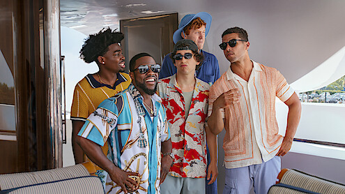 Five men in colorful summer shirts are standing and talking on a luxury boat deck, smiling and laughing together in a relaxed, sunny outdoor setting.
