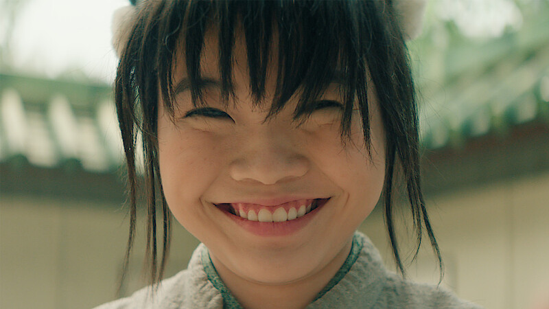 A smiling child with dark hair and bangs, wearing a light-colored top, stands outdoors in a courtyard with greenery and traditional architecture in the background.