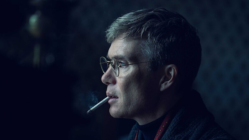 A man with short hair and glasses in a dark, softly lit room is shown in profile view, wearing a dark jacket and smoking a cigarette, smoke rising in front of his face, with a blurred, patterned background.