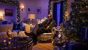 Man in a suit sits on a luxurious chair in an elegant, festively decorated living room with Christmas tree, gifts, candles, and warm lighting, creating a cozy and celebratory mood.