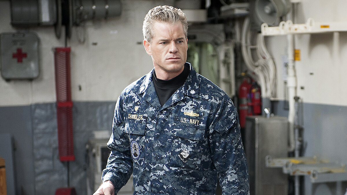 A man in a blue digital camouflage Navy uniform stands indoors on a ship, surrounded by industrial equipment, pipes, and emergency gear, looking serious and focused.