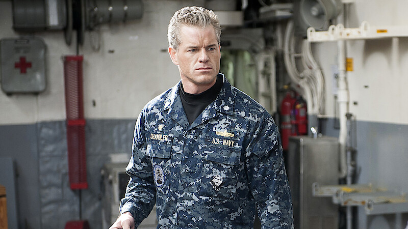 A man in a blue digital camouflage Navy uniform stands indoors on a ship, surrounded by industrial equipment, pipes, and emergency gear, looking serious and focused.