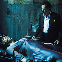 A man in a tuxedo stands beside a restrained woman lying on a table in a dim, grimy room with medical equipment and worn blue-green walls, suggesting a tense or dramatic scene.