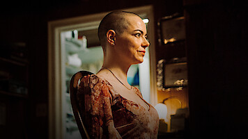 A woman with a shaved head sits in profile in a warmly lit room, smiling. The background includes a doorway revealing a bright, cluttered space and certificates on the wall, suggesting a personal or home environment.
