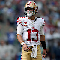 Football player wearing a San Francisco 49ers uniform and helmet stands on the field during a game, looking to the side, with a blurred stadium crowd in the background.