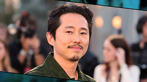 Steven Yeun with short dark hair and trimmed beard wearing a green jacket, standing outdoors at an event with blurred people, cameras, and lights in the background, creating a professional and lively atmosphere.