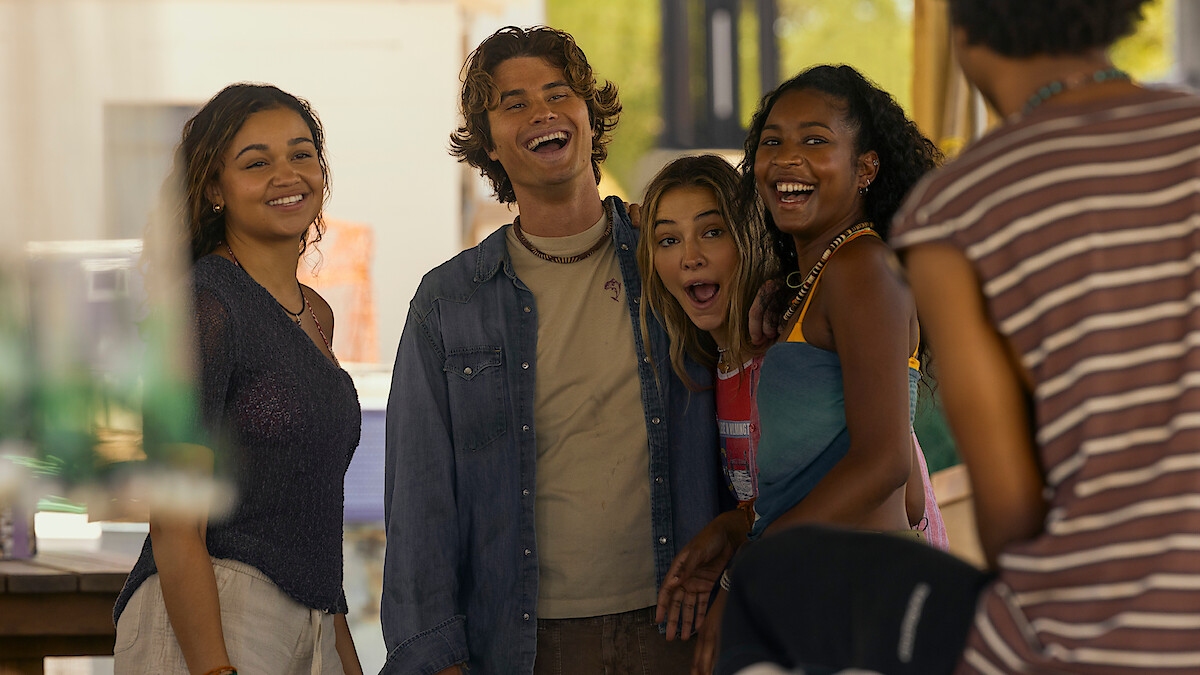 Four young adults stand closely together, smiling and laughing outdoors in a casual, sunny setting, with another person partially visible in the foreground. The group appears to be enjoying a relaxed, friendly moment together.