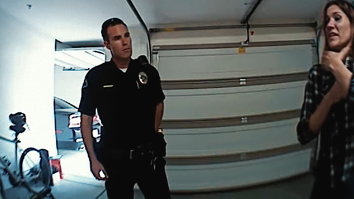 A police officer stands in a garage near a woman who appears to be explaining something. There is a bicycle by the garage door, and the setting looks like a residential area.