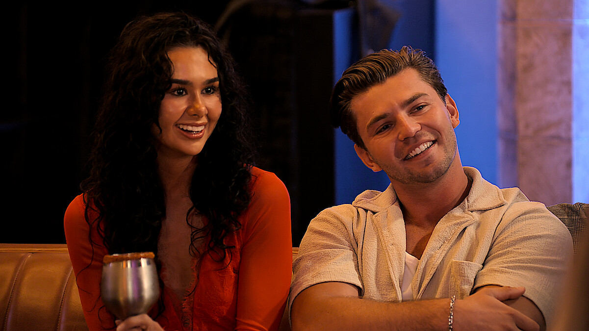 Two young adults sitting on a couch indoors, smiling and looking toward someone off camera. The woman holds a drink, both appear relaxed. Warm lighting suggests a social, casual atmosphere.