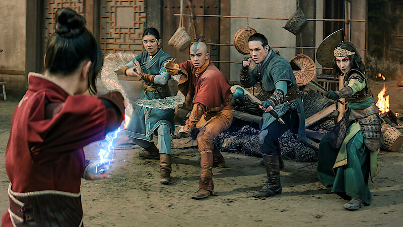 Four young warriors in fantasy costumes face an opponent producing blue energy in an ancient, dimly lit building with oriental decor and scattered debris, suggesting a tense magical battle scene.