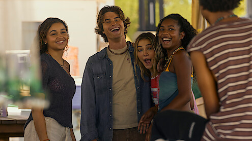 Four young adults stand closely together, smiling and laughing outdoors in a casual, sunny setting, with another person partially visible in the foreground. The group appears to be enjoying a relaxed, friendly moment together.