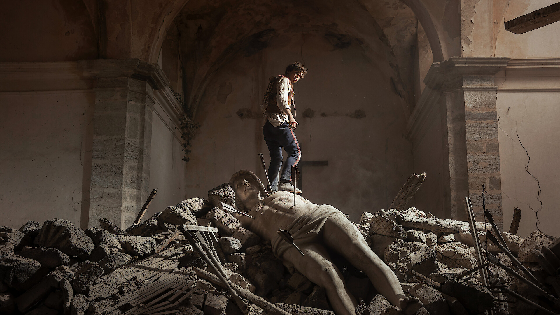 A person standing on rubble inside a damaged, old stone building, with a large fallen classical statue lying among the debris under an arched ceiling, evoking a sense of destruction and loss.