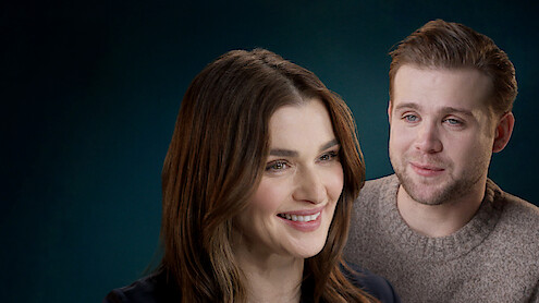 Two people indoors, a woman in the foreground smiling and a man in the background with a neutral expression, both against a dark, softly lit background.
