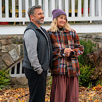 Man and woman standing outdoors in autumn leaves, in front of a white fence and stone wall, both dressed warmly in fall clothing; the woman wears a purple beanie and plaid jacket, the man is smiling, casual setting.