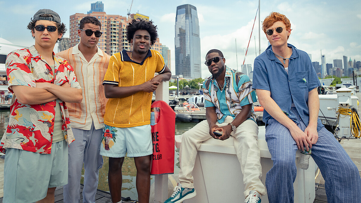 Five young men in casual summer clothes pose by a marina with boats, modern high-rise buildings, and the city skyline visible in the background on a sunny day.