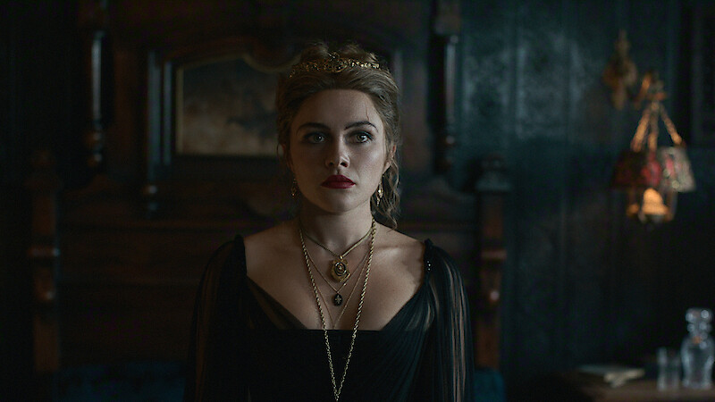 A woman in a dark, ornate dress with layered necklaces stands in a dimly lit, elegant room with vintage wooden furniture and decorative lighting, creating a moody and dramatic atmosphere.