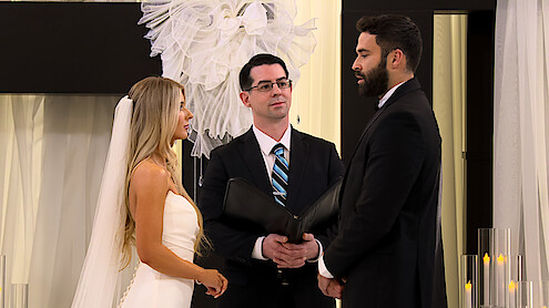 Ashley Carpenter and Alex Henderson stand facing each other in a formal indoor wedding ceremony with an officiant between them, surrounded by white drapery and candles.