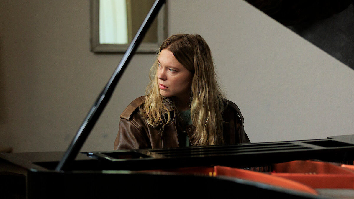 A person with long hair and a brown jacket sits at a grand piano indoors, looking to the side. The setting is softly lit with a neutral wall and a mirror in the background.
