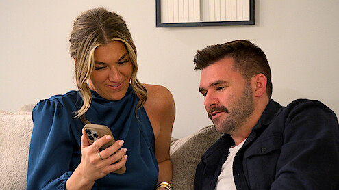 Connor and Brianna sitting on a couch indoors, smiling while looking at a smartphone together, casual home setting with light-colored walls and framed art in the background.
