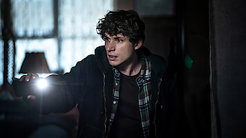 A young man in casual clothes cautiously searches a dimly lit, cluttered room with a flashlight, appearing tense and alert in a possibly abandoned or mysterious setting.