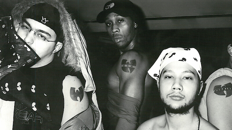Three men pose in a black and white indoor photo, showing off Wu-Tang Clan tattoos and clothing featuring the group's logo. The environment is casual with urban streetwear and bandanas, emphasizing their affiliation and attitude.