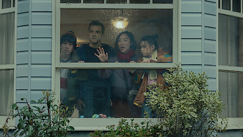 Four people stand closely together indoors, looking anxiously out a rainy window of a house with pale siding, surrounded by green shrubs, under soft indoor lighting.