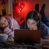 Two young women lying on a bed in a cozy, dimly lit room with posters and string lights, watching something on a laptop with a bowl of popcorn nearby.