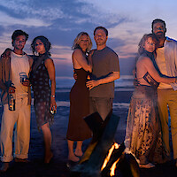 Six adults standing in three couples hug around a beach bonfire at sunset, with the ocean and sky in the background. They appear relaxed and intimate, holding drinks and wearing summer evening clothing.