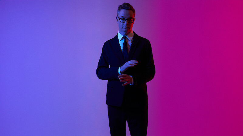 Nicolas Winding Refn wears a black suit, white shirt, and dark tie, and adjusts his cuffs against a pink and purple neon background. 