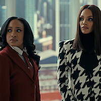 Two women in professional outfits stand indoors with a cityscape visible through glass windows behind them, appearing serious and focused in a modern office setting.